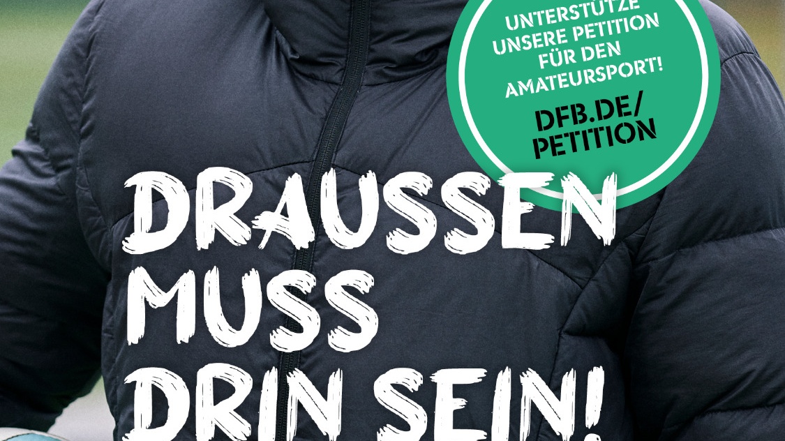 Mann mit blonden Haaren auf Fußballfeld, trägt Trainingsjacke mit Aufschrift "DRAUSSEN MUSS DRIN SEIN!" und Petition für Amateursport. Im Hintergrund grüner Rasen.