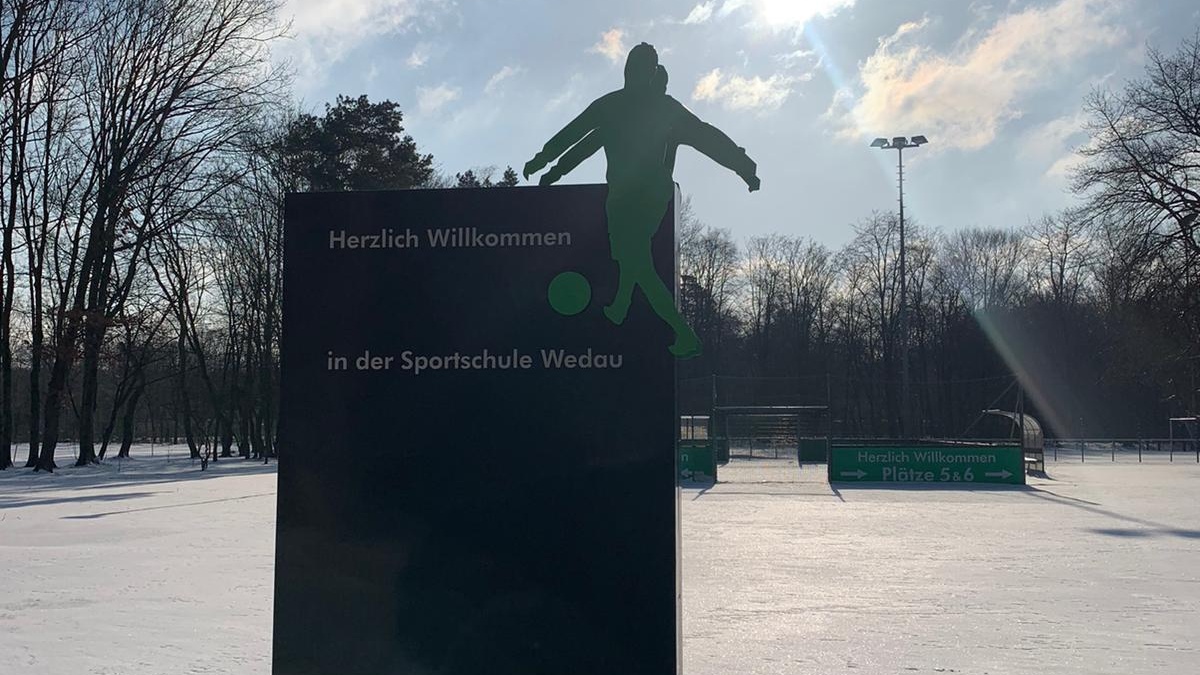Schneebedeckter Sportplatz mit Fußballer-Silhouette auf einem schwarzen Wegweiser. Im Hintergrund kahlste Bäume und ein hellblauer Himmel mit Sonne.