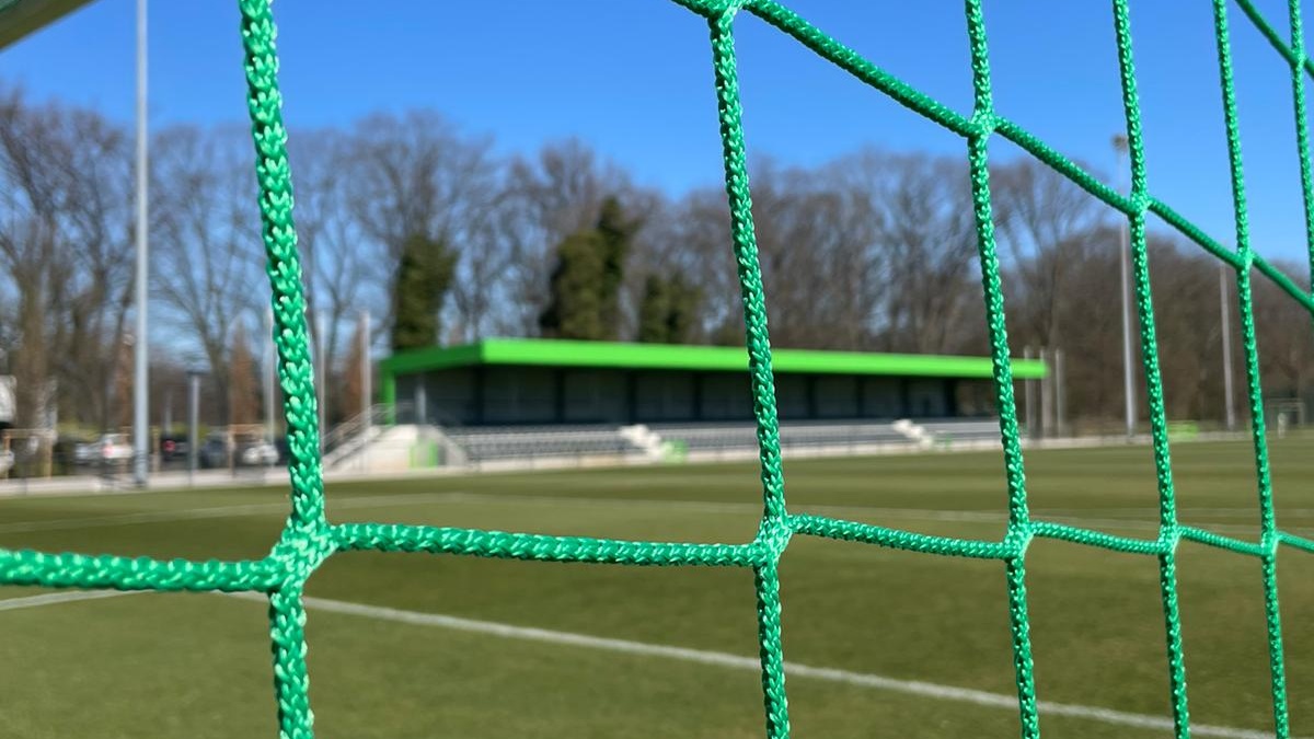 Fußballtor mit Netz, Blick auf grünen Rasen und Fußballfeld. Im Hintergrund Gebäude und Bäume unter blauem Himmel.
