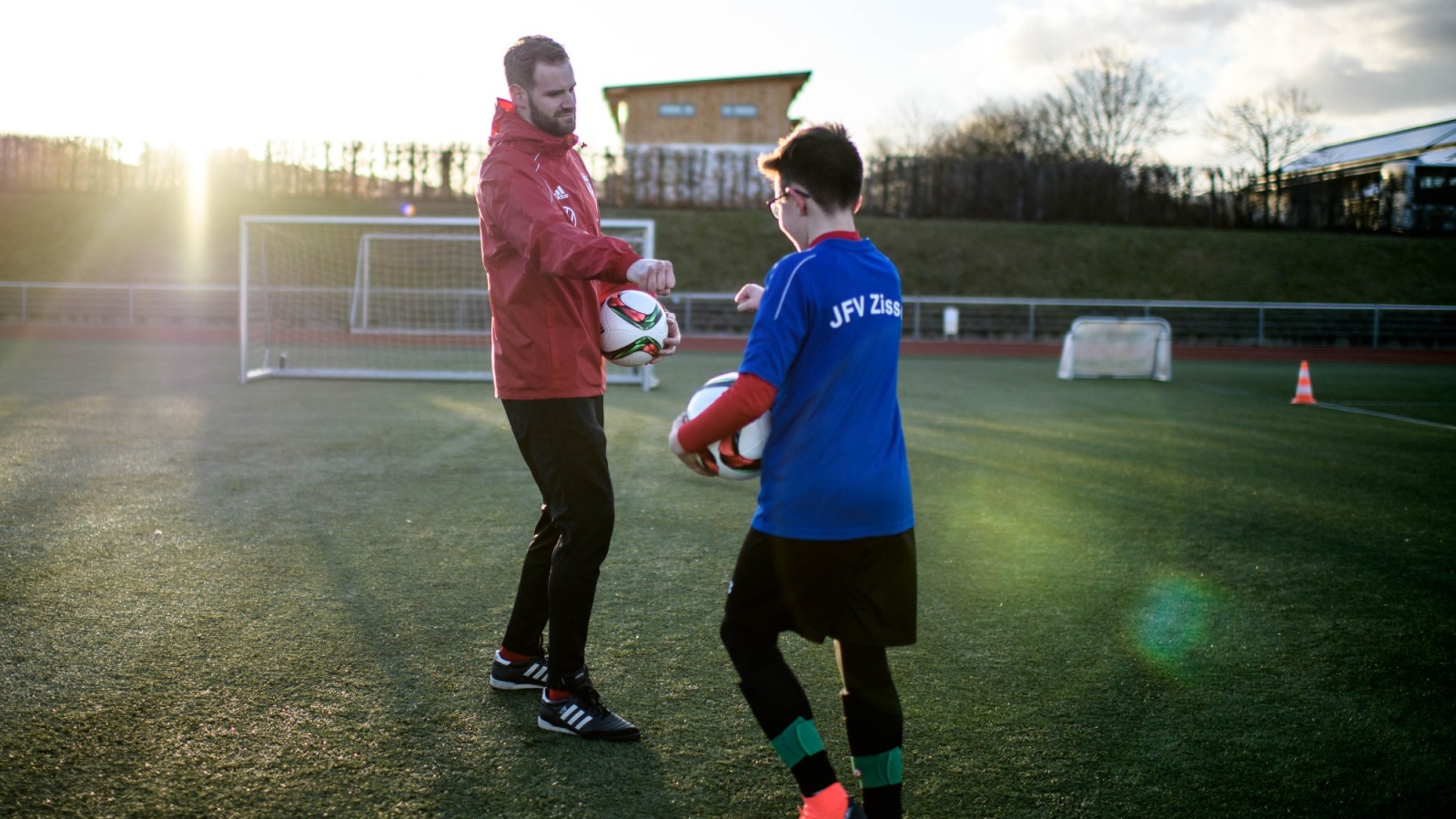 Zwei Personen auf einem Fußballfeld: Ein Mann in roter Jacke erklärt einem Jungen mit blauer Sportbekleidung etwas und hält einen Fußball.