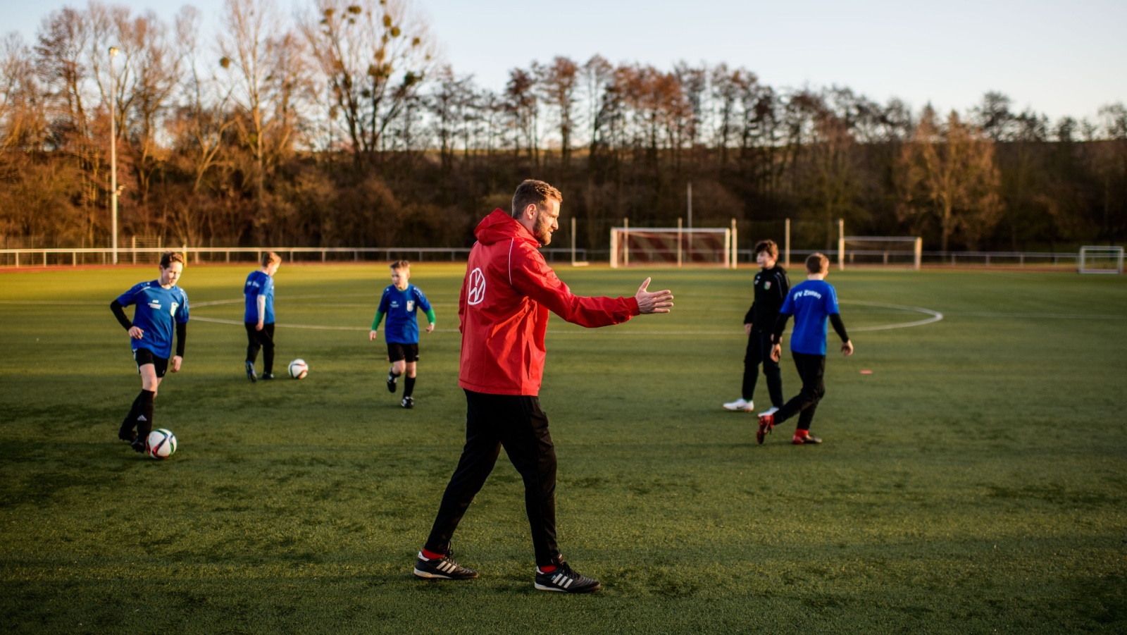 Fußballtrainer mit roter Jacke auf grünem Kunstrasenplatz. Mehrere Kinder in blauem Trikot trainieren mit Ball. Im Hintergrund Bäume und Fußballtor.