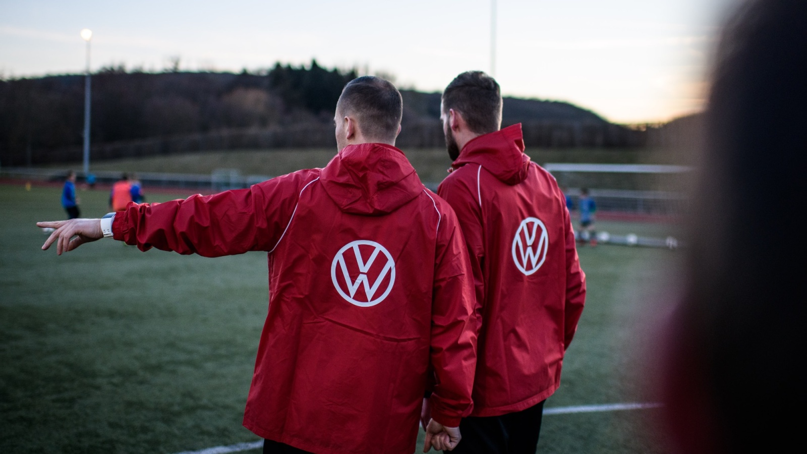 Zwei Männer mit roten Jacken und VW-Logo stehen auf einem Fußballfeld und weisen in Richtung Spielfeld. Im Hintergrund weitere Spieler und Bäume.