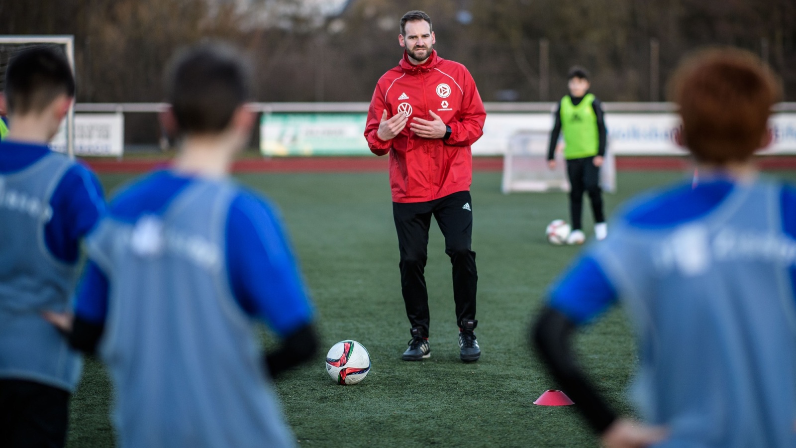 Fußballtrainer mit roter Jacke vor Jugendmannschaft auf grünem Kunstrasen. Spieler tragen blaue Trainingswesten, Fokus auf den Ball vor dem Trainer.