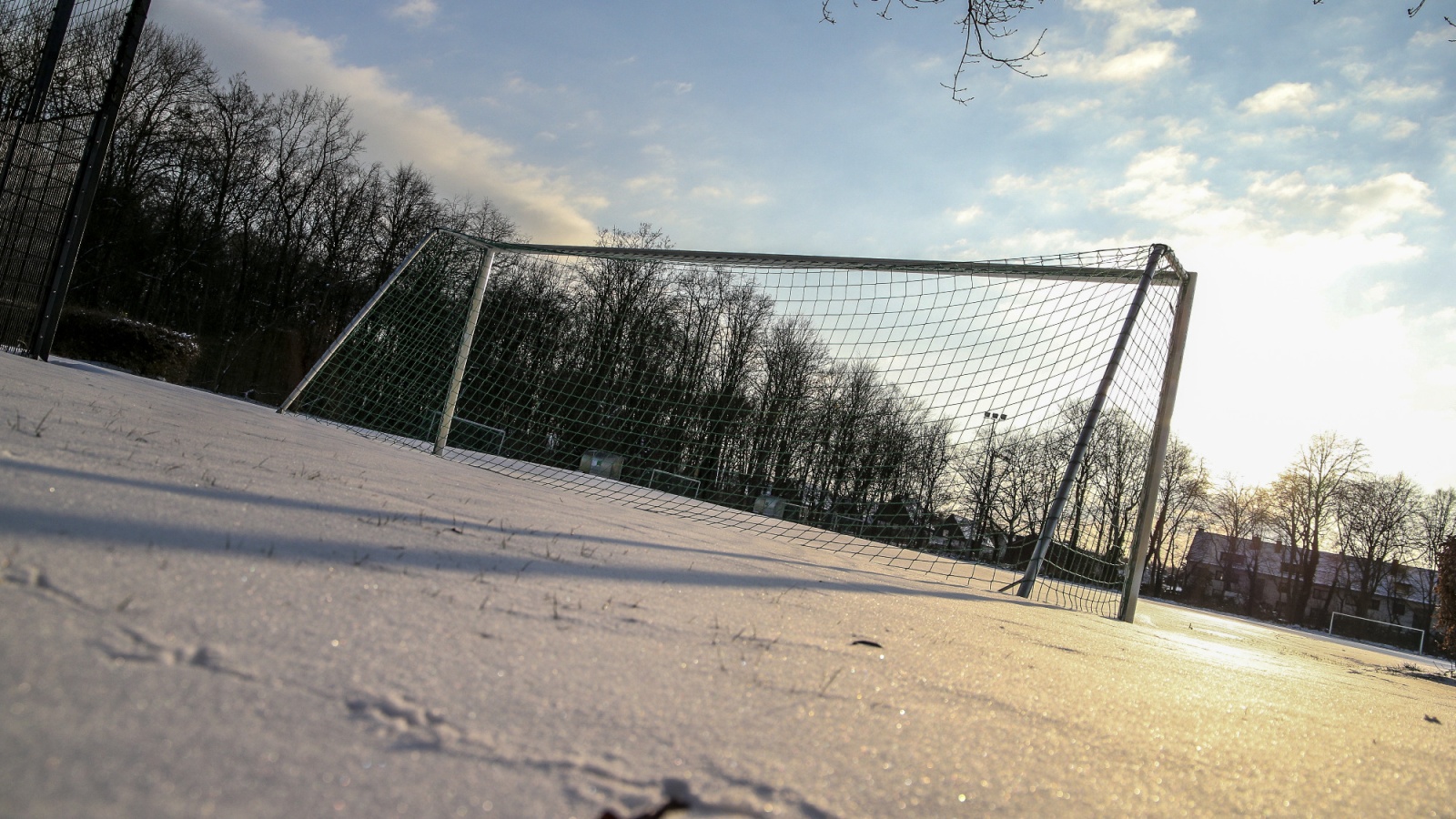 Schneebedeckter Fußballplatz. Im Vordergrund das Fußballtor mit Netz. Im Hintergrund Bäume und Gebäude unter winterlichem Himmel.