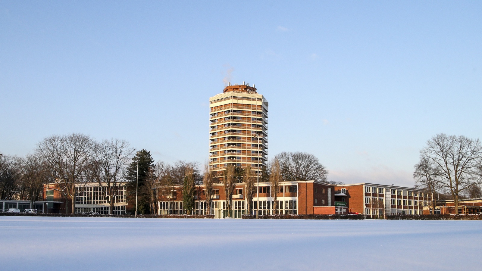 Hoher Wohnturm mit Flachdach, davor flache Gebäude mit langen Reihenfenstern. Im Vordergrund schneebedeckte Fläche, dahinter kahle Bäume und klarer Himmel.
