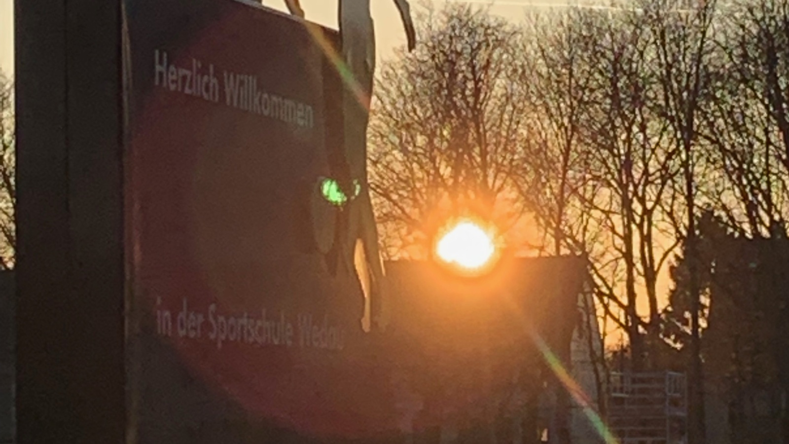 Schild mit Silhouetten von Personen, die Hände halten, mit dem Text "Herzlich Willkommen in der Sportschule Wedau". Dahinter geht die Sonne auf, Bäume im Hintergrund.