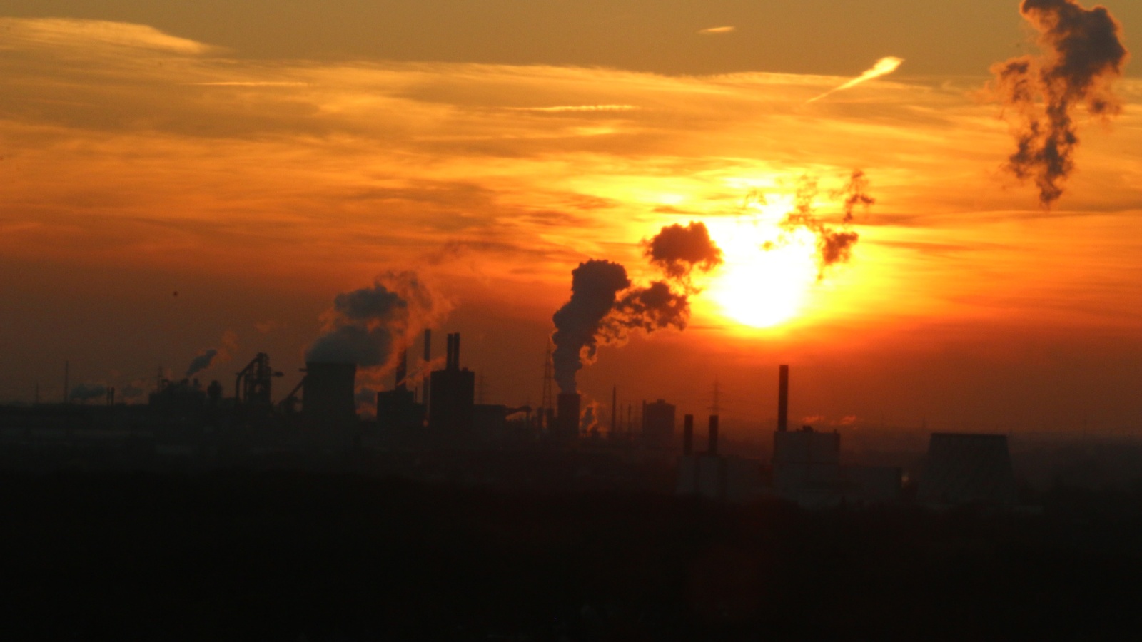 Industrielandschaft mit Fabriken und Schloten im warmen Licht einer tiefstehenden Sonne. Rauch steigt auf.