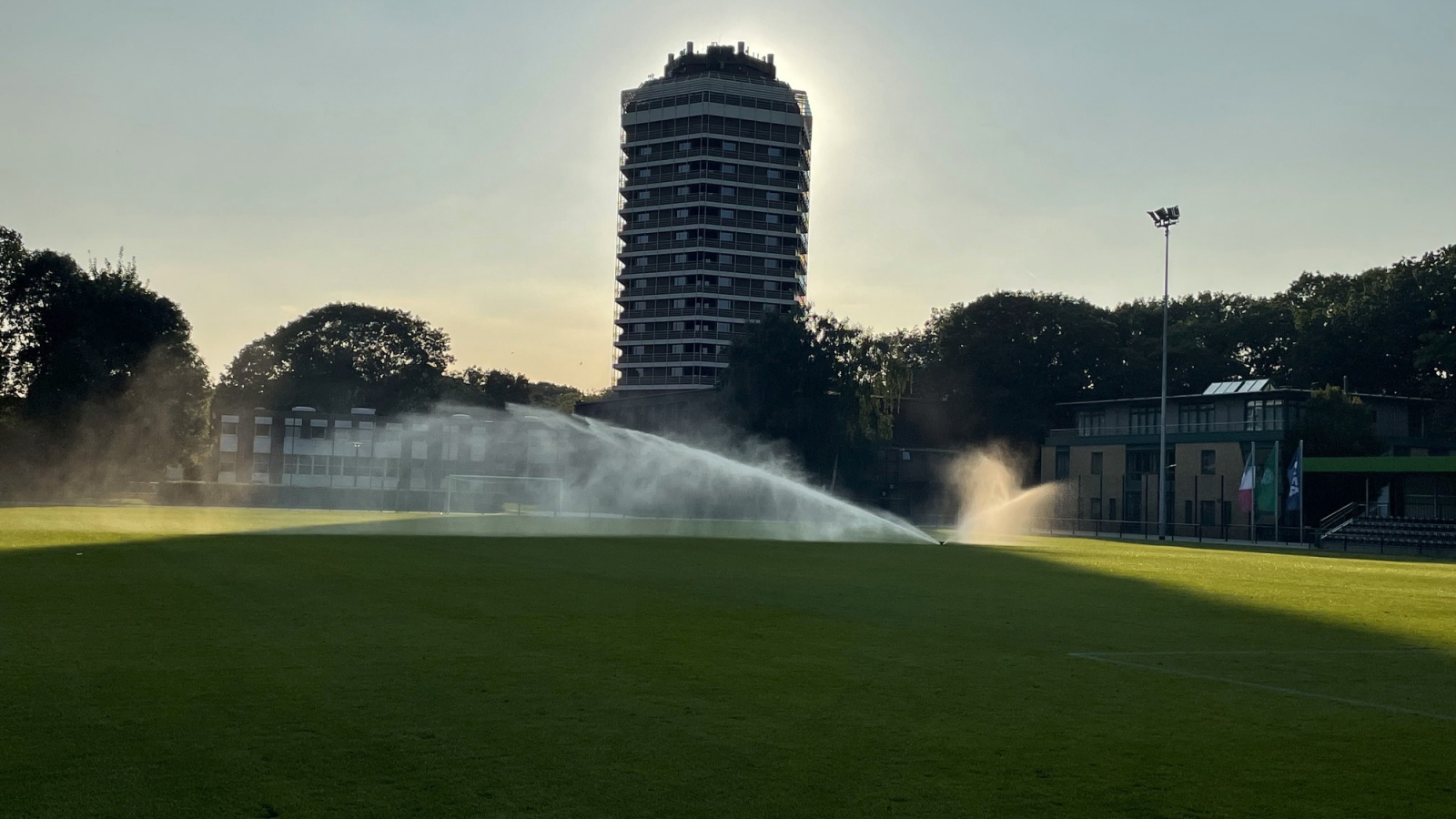 Grüner Rasenplatz mit Sprühnebeln durch Bewässerungsanlage. Im Hintergrund ein hoher Wohnblock und Bäume unter hellem Himmel.