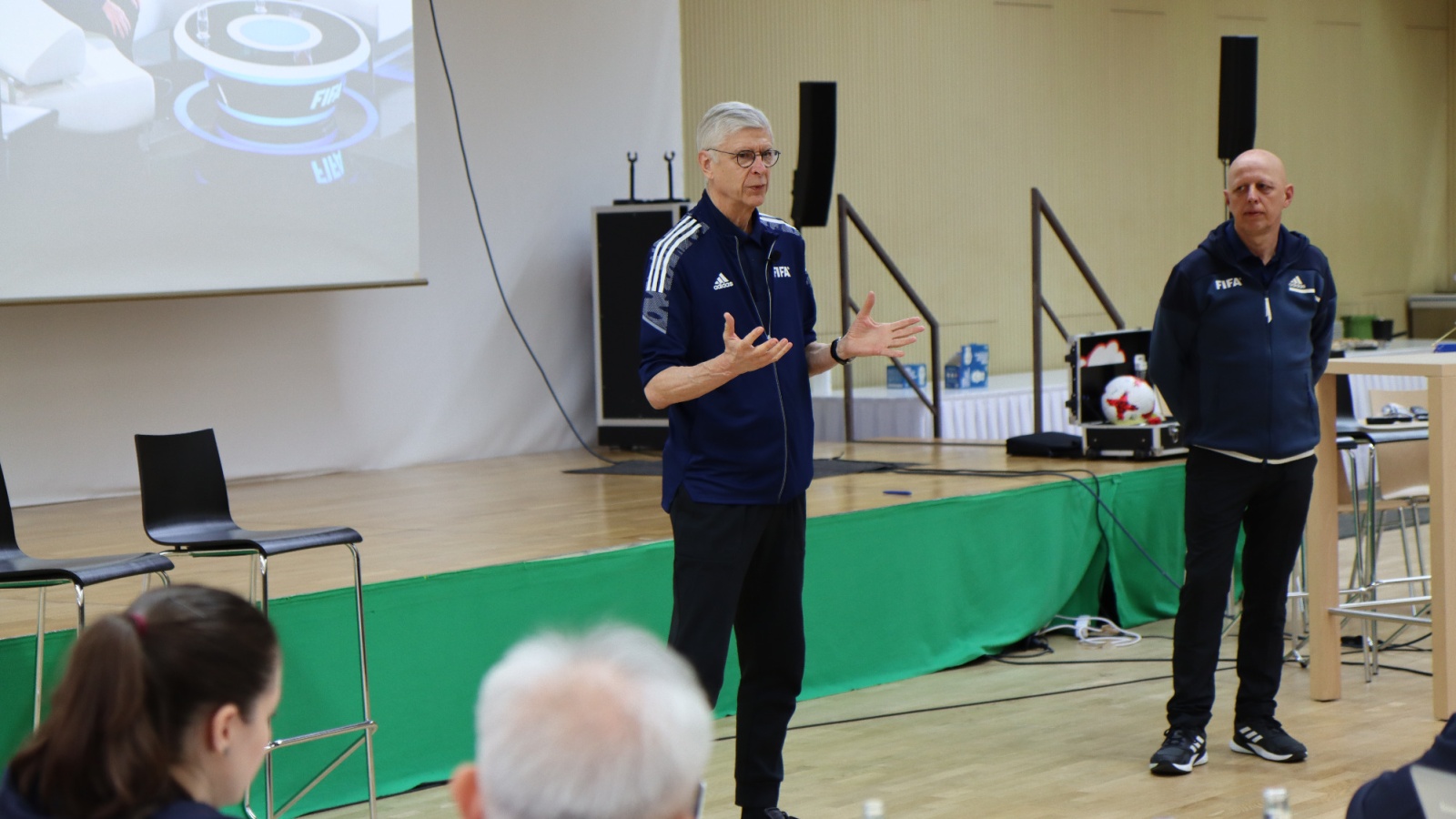 Zwei Männer in Trainingsanzügen stehen vor einer Projektion und sprechen. Im Publikum sitzen Personen an einem Tisch. Konferenzraum mit Parkettboden.