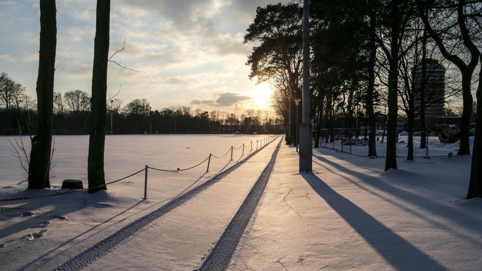 Schnee bedeckter Parkweg mit Bäumen und im Hintergrund ein zugefrorener See bei Sonnenuntergang. Lange Schatten fallen auf den Schnee.