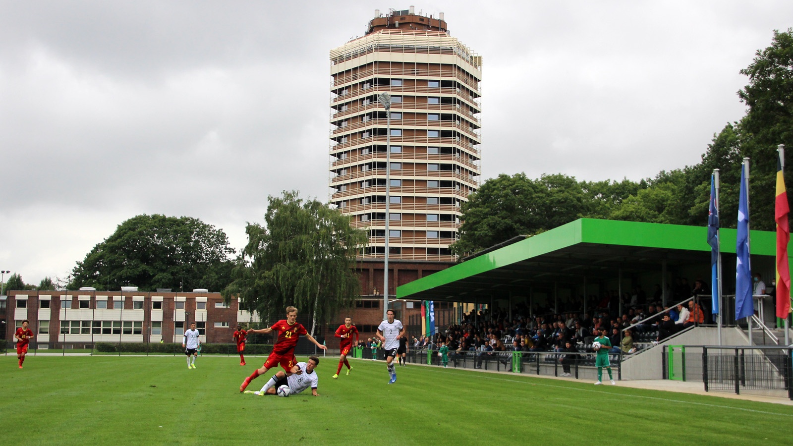 Fußballspiel im Stadion. Spieler in rot und schwarz auf grünem Rasen. Im Hintergrund ein hoher Wohnturm und Zuschauerüberdachung.