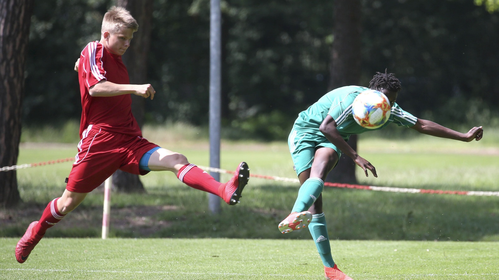 Zwei jugendliche Fußballspieler auf Grün. Links ein Spieler in Rot, rechts in Grün, beim Schuss auf den Ball. Hintergrund: Bäume.