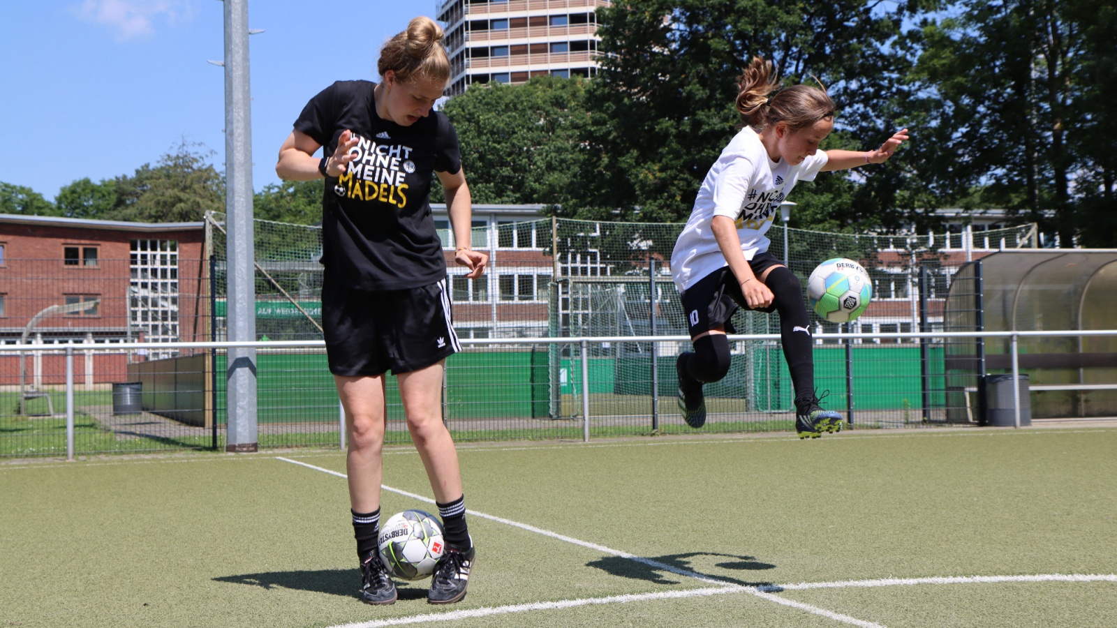 Zwei jugendliche Fußballspielerinnen trainieren auf einem grünen Kunstrasenplatz. Eine jongliert mit dem Ball, die andere schießt. Im Hintergrund Gebäude und ein Zaun.