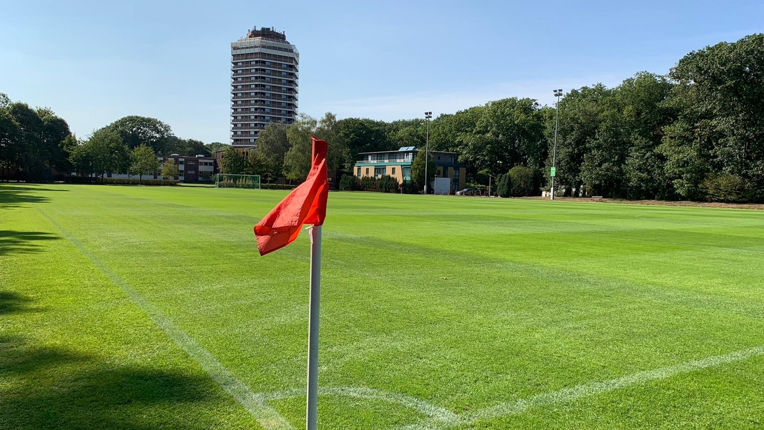 Fußballfeld mit roter Eckeinwurf-Flagge. Im Hintergrund ein Hochhaus und Bäume unter blauem Himmel.