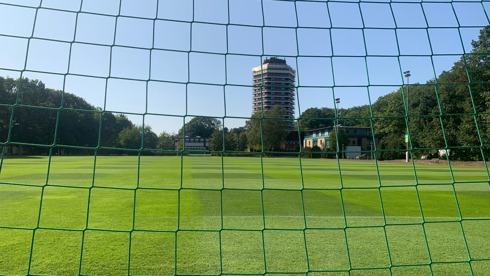 Fußballfeld mit grüner Netztür im Vordergrund. Im Hintergrund Gebäude und Bäume unter blauem Himmel.