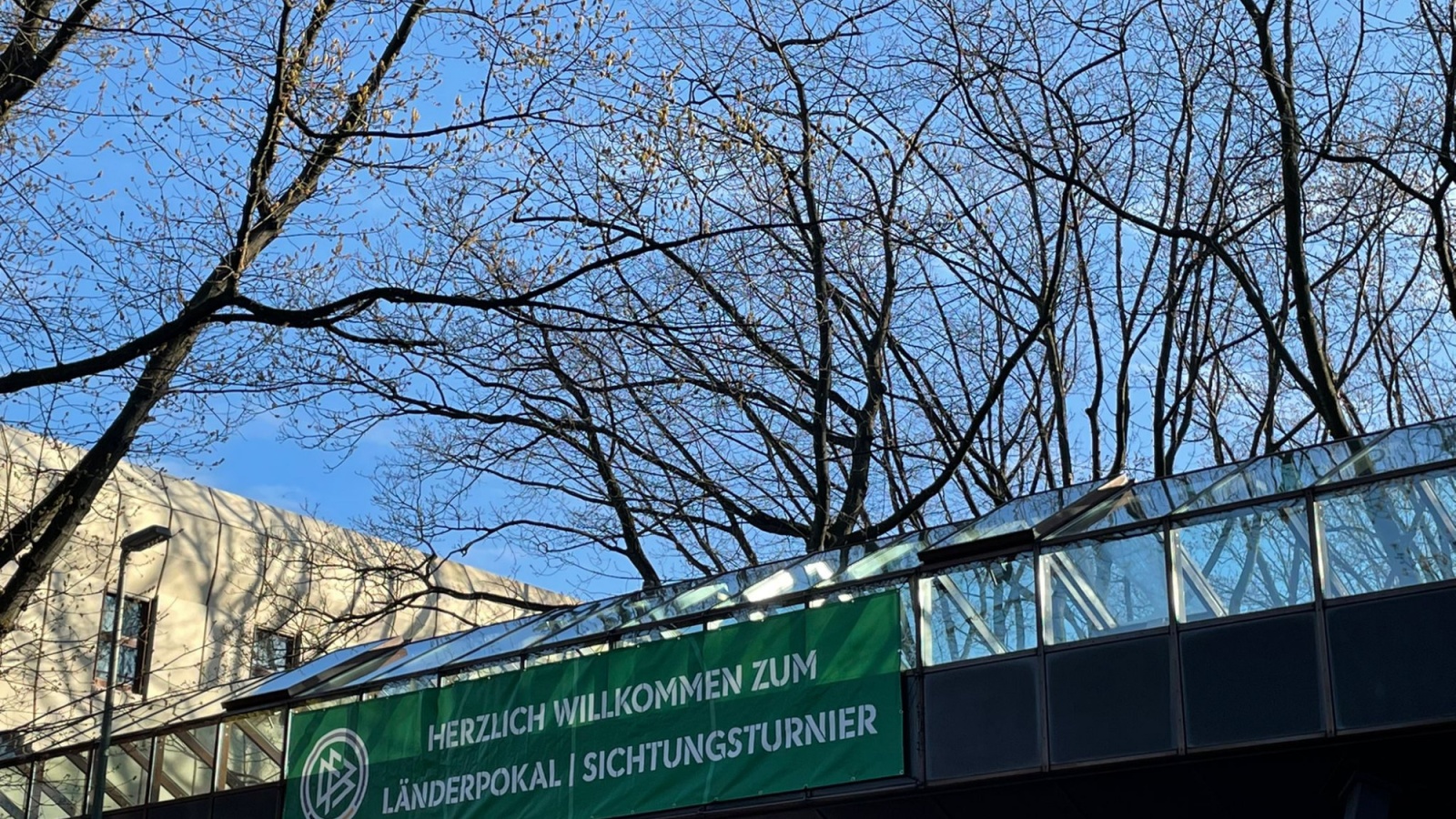 Überdachung eines Sportgeländes mit Banner "HERZLICH WILLKOMMEN ZUM LÄNDERPOKAL | SICHTUNGSTURNIER". Dahinter Bäume gegen blauen Himmel.