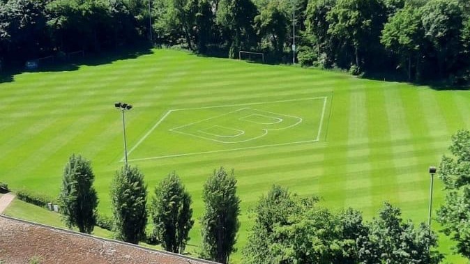 Luftaufnahme einer grünen Sportwiese mit einem weißen Logo in der Mitte, umgeben von Bäumen und einem Gebäude mit Satteldach im Vordergrund.