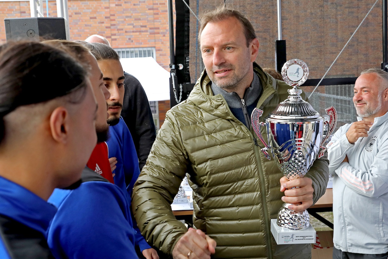 [{"clang_id":1,"value":"Mann mittleren Alters mit Jacke hält Pokal, schüttelt Hände mit zwei jungen Fußballspielern. Im Hintergrund weitere Personen und eine Backsteinmauer."}]