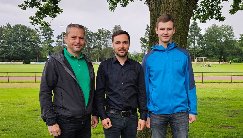 [{"clang_id":1,"value":"Drei Männer stehen nebeneinander auf einer grünen Wiese vor einem Fußballfeld und Bäumen. Links trägt einer eine schwarze Jacke und grünes Hemd, in der Mitte einen schwarzen Blusen und rechts einen blauen Hoodie."}]