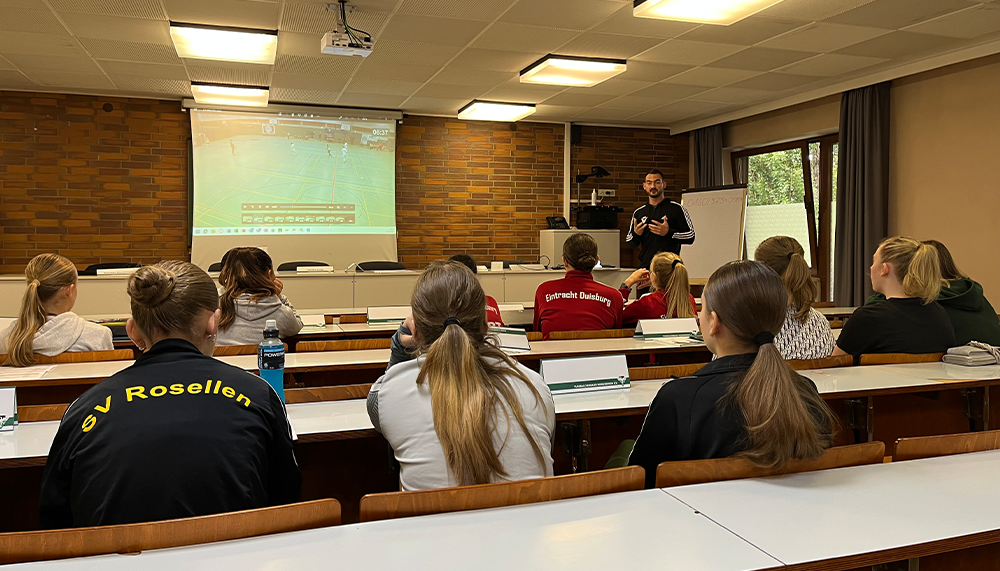 [{"clang_id":1,"value":"Klassenzimmer mit Schülern, die einem Lehrer zuhören. An der Wand ein Beamer mit Fußballfeld-Darstellung. Einige Schüler tragen Sportkleidung."}]