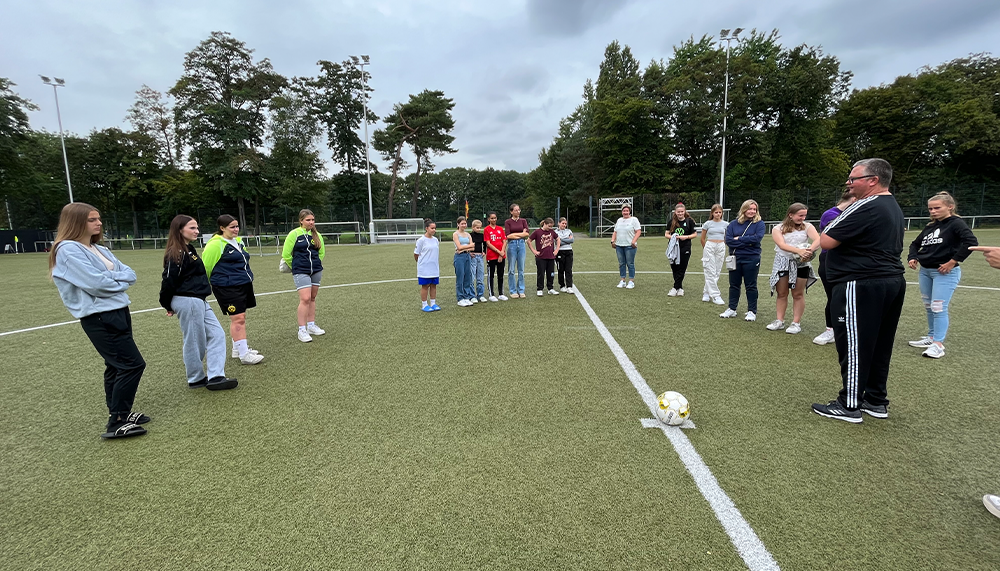 [{"clang_id":1,"value":"Fußballfeld mit Trainer und etwa 20 Kindern/Jugendlichen im Kreis. Alle schauen auf einen Fußball in der Feldmitte. Bäume im Hintergrund."}]