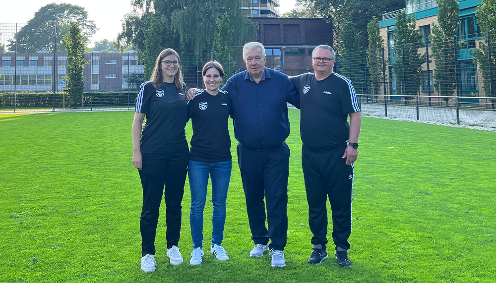 [{"clang_id":1,"value":"Vier Personen stehen auf einem Fußballfeld vor Gebäuden. Sie tragen Sportkleidung mit einem Logo. Von links: Frau, Frau, Mann, Mann."}]