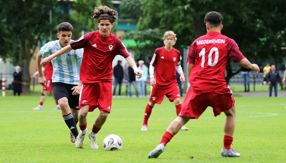 [{"clang_id":1,"value":"Jugendfußballspiel. Drei Spieler in roten Trikots und ein Spieler in blau-weißem Trikot kämpfen um den Ball auf einem Rasenplatz. Im Hintergrund Zuschauer und Bäume."}]