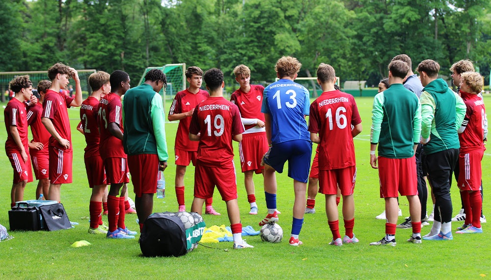 [{"clang_id":1,"value":"Fußballmannschaft im Kreis auf dem Rasen. Spieler tragen rote und blaue Trikots, einige stehen am Rand. Im Hintergrund grüne Bäume und Fußballtor."}]