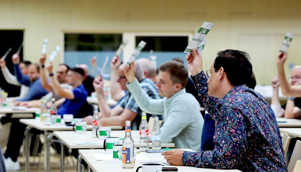 [{"clang_id":1,"value":"Versammlung in einem Konferenzraum. Teilnehmer heben grüne und weiße Karten in die Luft. Einige tragen Hemden, andere dunkle Jacken. Tische mit Wasserflaschen."}]
