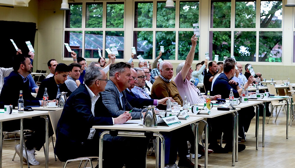 [{"clang_id":1,"value":"Versammlung von Männern in Anzügen an Tischen, viele heben grüne Karten hoch. Fenster im Hintergrund zeigen Bäume."}]