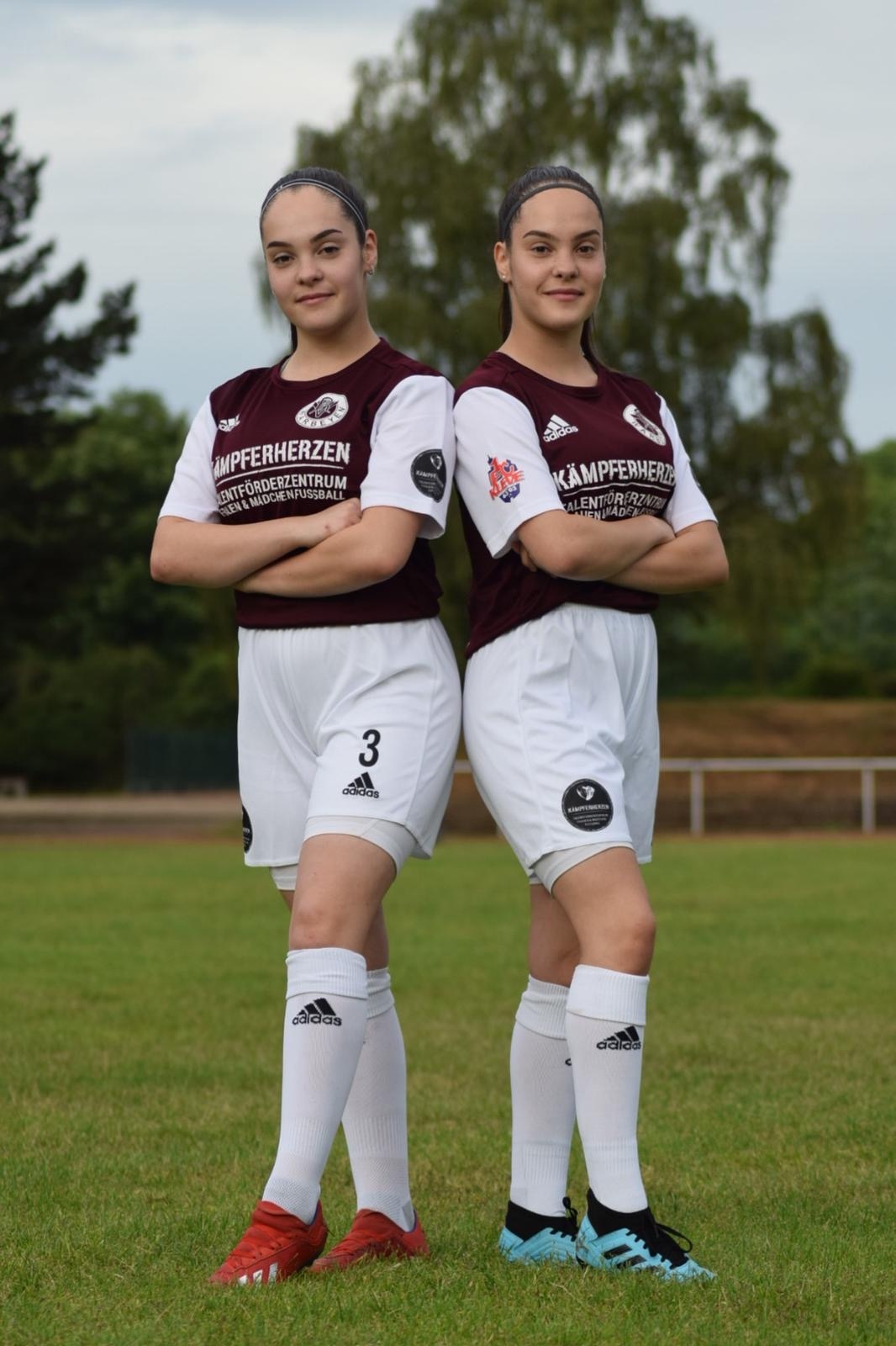 [{"clang_id":1,"value":"Zwei junge Fußballspielerinnen im Vereinstrikot (maroon/weiß) stehen auf einem Rasenplatz. Beide tragen Sportschuhe und weiße Socken."}]