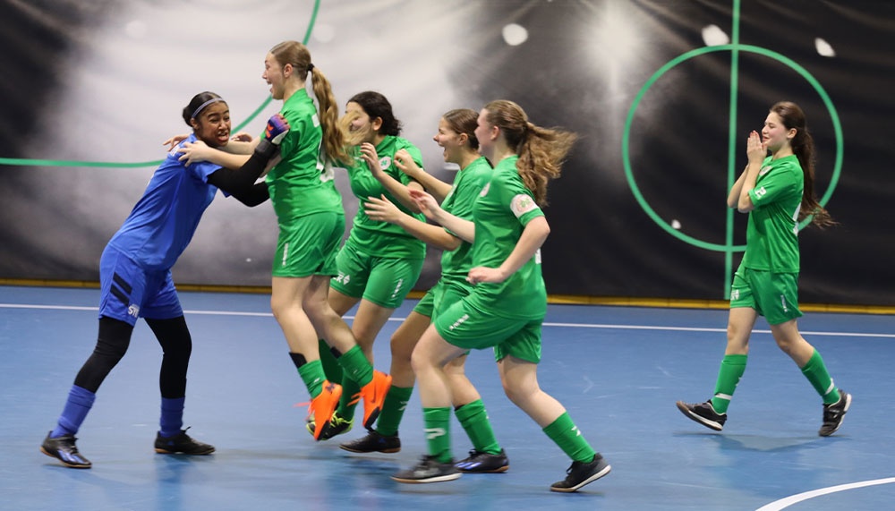 [{"clang_id":1,"value":"Futsal-Spiel: Gruppe jubelnder Spielerinnen in grünen Trikots, eine Spielerin in blauem Trikot hält eine jubelnde Spielerin fest. Hallenfußballfeld."}]