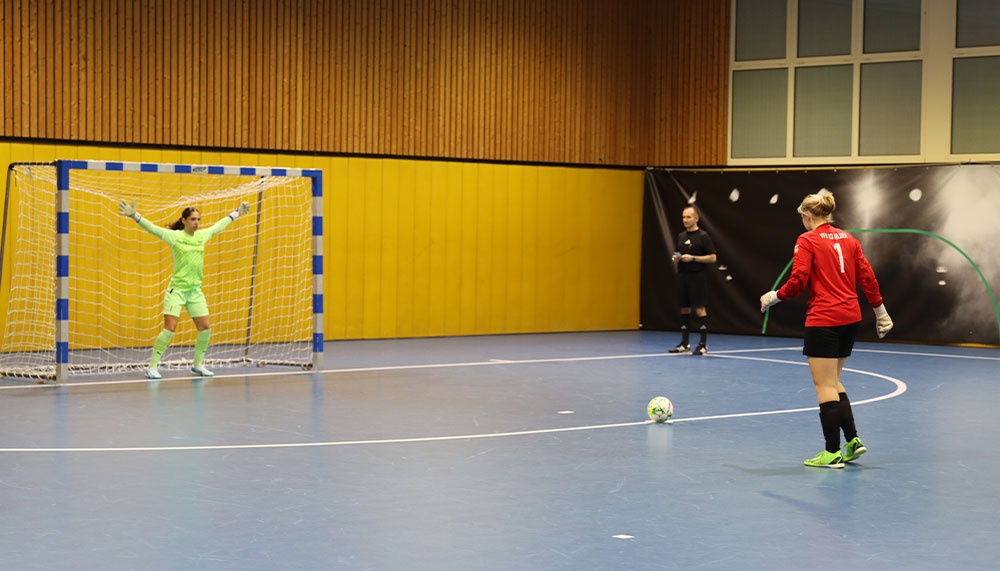 [{"clang_id":1,"value":"Futsalspiel: Torwartin in grün vor dem Tor, Spielerin in rot mit Ball, Schiedsrichter im Hintergrund. Sporthalle mit Holzvertäfelung."}]