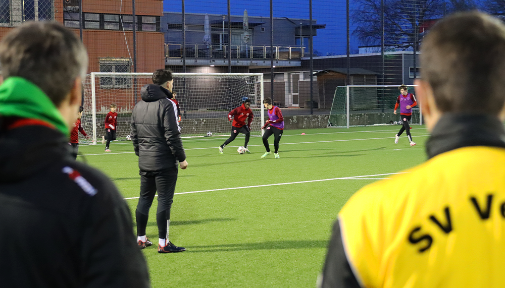 [{"clang_id":1,"value":"Fußballtraining auf Kunstrasenplatz. Spieler in Rot-Lila, Trainer beobachten. Im Hintergrund Gebäude und Zaun. Vordergrund verschwommen mit gelber Weste und grüner Jacke."}]