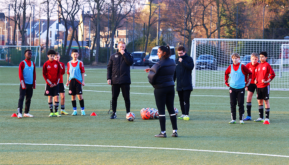 [{"clang_id":1,"value":"Fußballtrainer und Jugendmannschaft auf grünem Kunstrasenplatz. Trainer erklären Taktik, Spieler stehen im Kreis. Im Hintergrund Gebäude und Bäume."}]