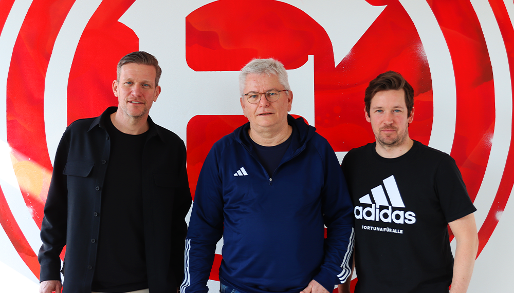 [{"clang_id":1,"value":"Drei Männer stehen vor einer Wand mit rotem Kreis und weißen Linien. Links trägt ein Mann dunkle Kleidung, in der Mitte ein blauen Trainingsanzug und rechts ein schwarzes T-Shirt mit weißem Adidas-Logo."}]