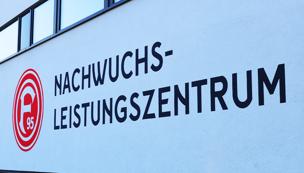 [{"clang_id":1,"value":"Gebäude mit Schriftzug \"NACHWUCHS-LEISTUNGSZENTRUM\" und Logo mit rotem Kreis, darin \"95\" und stilisierter Helm. Hellblaue Fassade, dunkler Dachrand."}]