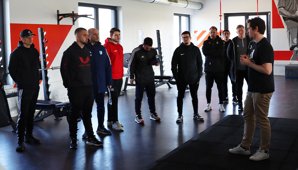 [{"clang_id":1,"value":"Gruppe von Männern in einem Fitnessstudio lauscht einem Trainer. Alle stehen im Kreis auf einem dunklen Holzboden, Trainingsgeräte im Hintergrund."}]