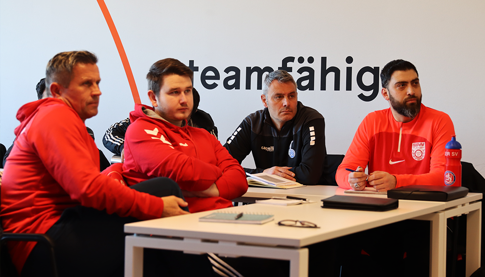 [{"clang_id":1,"value":"Vier Männer sitzen an einem Konferenztisch. Sie tragen Sportkleidung in Schwarz und Rot. Im Hintergrund ein weißes Banner mit dem Schriftzug \"teamfähig\"."}]
