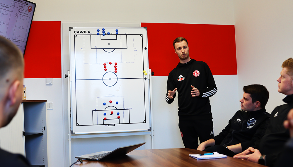 [{"clang_id":1,"value":"Fußballtrainer erklärt Taktik an einer Whiteboard-Tafel mit Spielfeldvorlage. Vier Spieler sitzen am Konferenztisch. Alle tragen Trainingsanzüge."}]