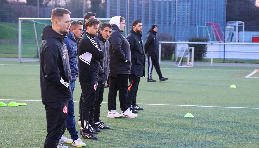[{"clang_id":1,"value":"Fußballmannschaft in Trainingskleidung steht an der Seitenlinie eines grünen Rasenplatzes. Im Hintergrund ein Zaun und Gebäude."}]