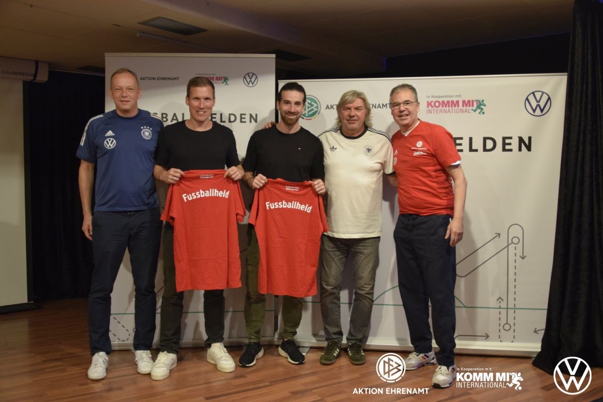 [{"clang_id":1,"value":"Fünf Männer stehen nebeneinander vor einem Banner mit Logos. Drei halten rote Fußballtrikots mit Aufschrift \"Fussballheld\" hoch."}]