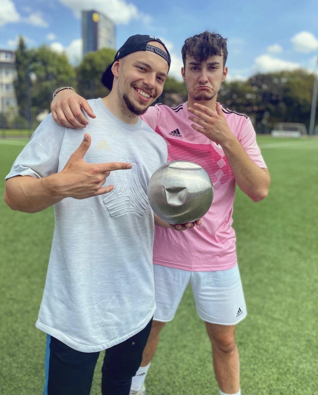 [{"clang_id":1,"value":"Zwei Männer auf grünem Rasen. Links lächelt ein Mann mit Baseballkappe und zeigt mit dem Finger nach unten. Rechts steht ein Mann in Sportkleidung, hält eine silberne Kuppel und blickt nachdenklich."}]