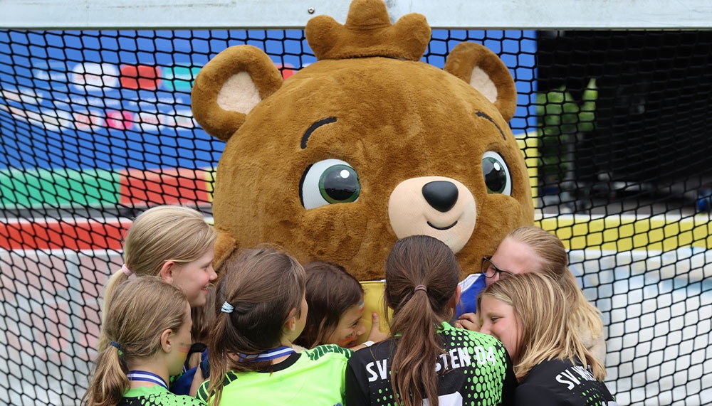 [{"clang_id":1,"value":"Fußballspielerinnen umarmen ein großes, braunes Bärenmaskottchen vor einem Fußballtor. Im Hintergrund ein farbiges Spielfeld."}]