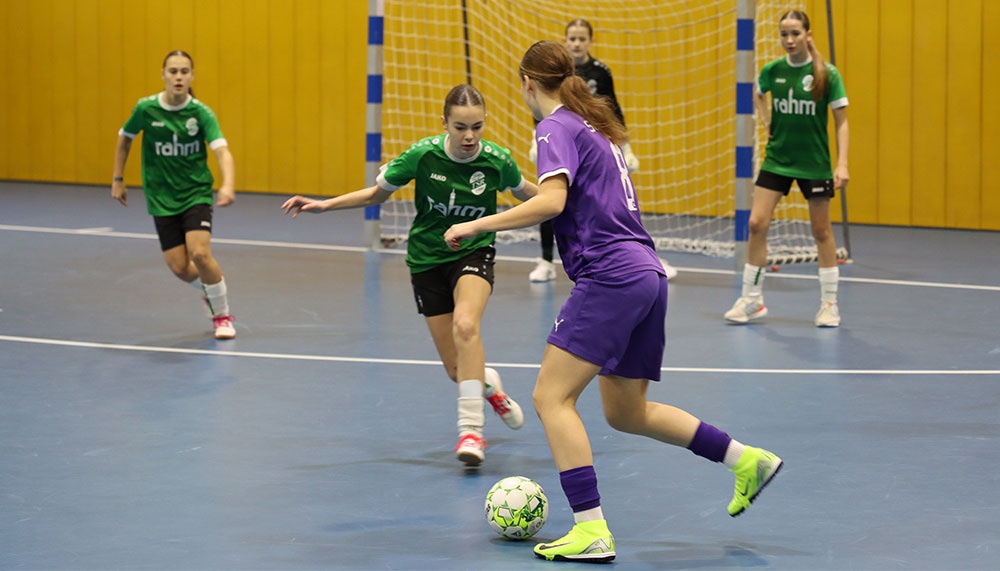 [{"clang_id":1,"value":"Futsalspiel in Sporthalle: Zwei Spielerinnen in Lila und Grün konzentriert am Ball, weitere Spielerinnen im Hintergrund. Netztor erkennbar."}]
