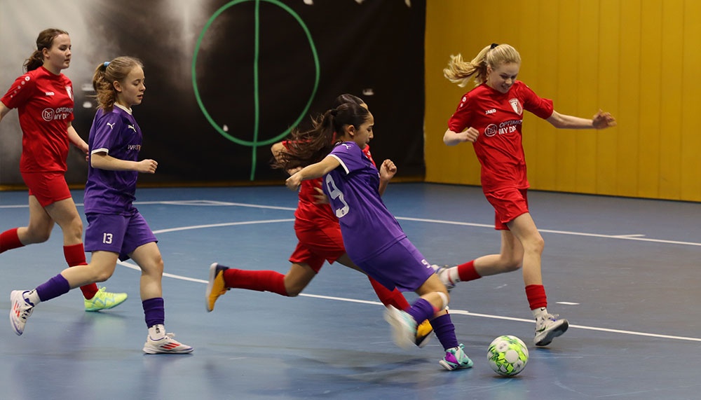 [{"clang_id":1,"value":"Fünf junge Fußballspielerinnen in Sporthalle. Zwei in Rot, zwei in Lila, eine in Blau. Eine Spielerin schießt auf den Ball."}]
