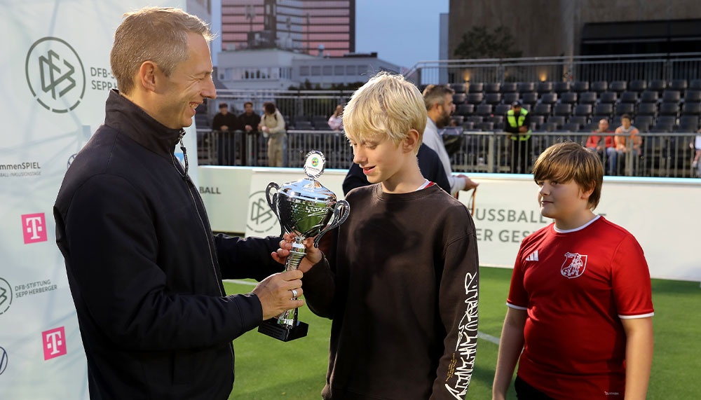 Mann übergibt Pokal an zwei Jungen auf Fußballfeld. Im Hintergrund Zuschauerränge und Werbebanner.