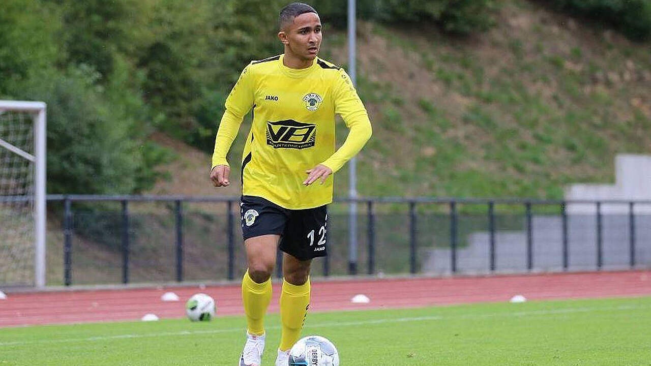 [{"clang_id":1,"value":"Fußballspieler mit gelbem Trikot und schwarzen Shorts auf grünem Rasen, dribbelt einen Ball. Im Hintergrund eine Zuschauertribüne und Bäume."}]