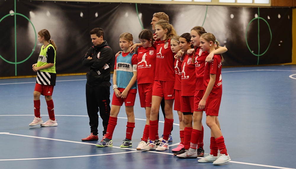 [{"clang_id":1,"value":"Handballteam mit Trainer in Sporthalle. Acht Mädchen in roten Trikots und ein Trainer in Schwarz stehen auf blauem Hallenboden. Eine Spielerin trägt eine gelbe Weste."}]