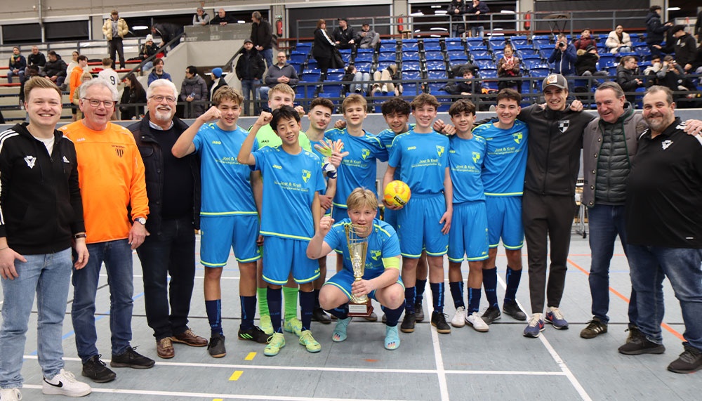 [{"clang_id":1,"value":"Jugendfußballmannschaft in blauen Trikots mit Trainern und Betreuern. Sie halten Pokal und Fußbälle in einer Sporthalle. Zuschauertribünen im Hintergrund."}]